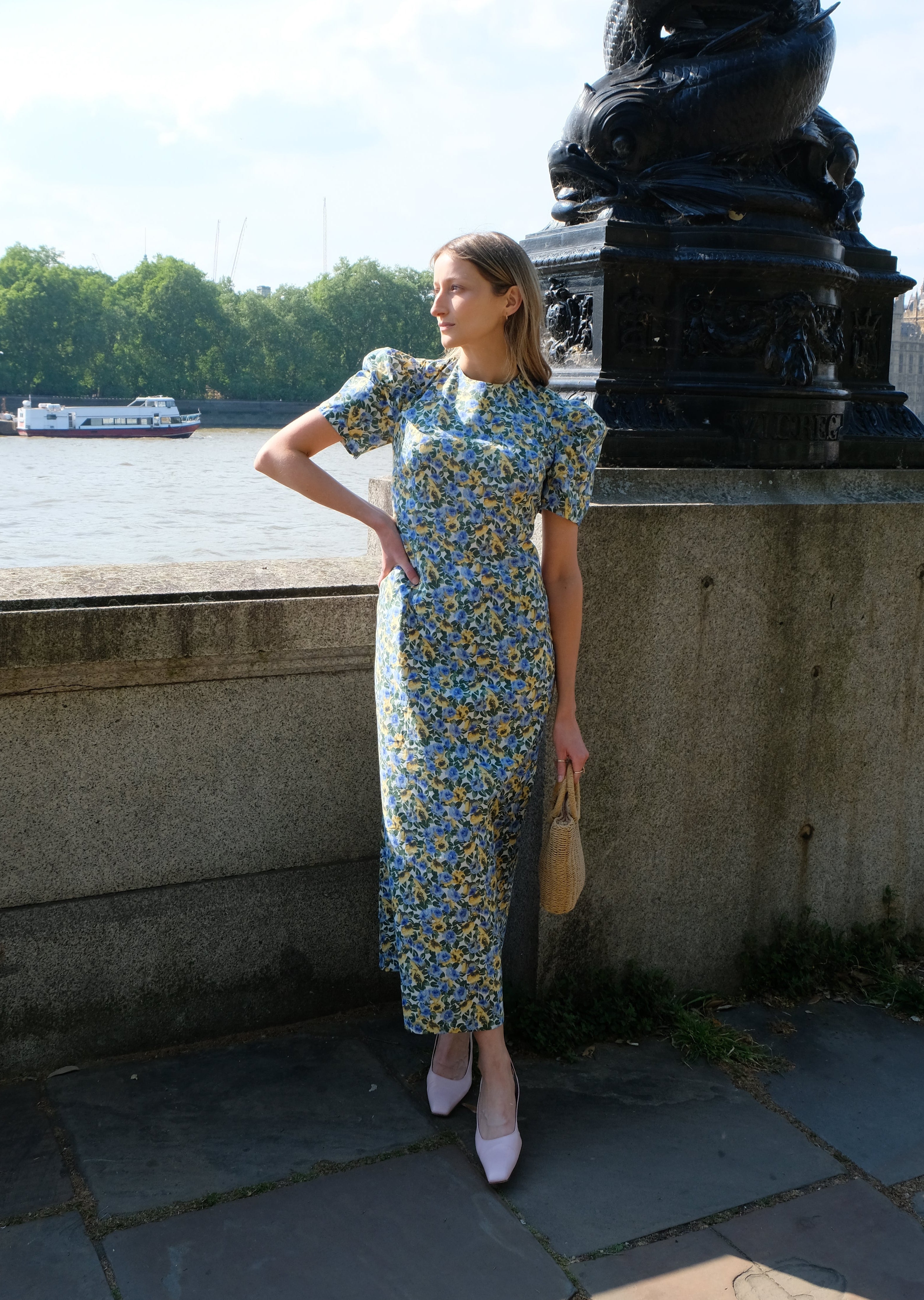 Woman in a floral dress standing by a river with a boat and city skyline in the background.